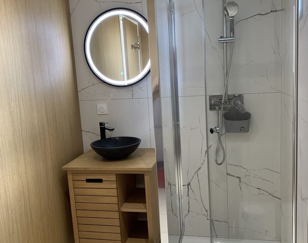 Salle de bain moderne dun hébergement insolite en Aquitaine, avec douche vitrée et miroir rond.