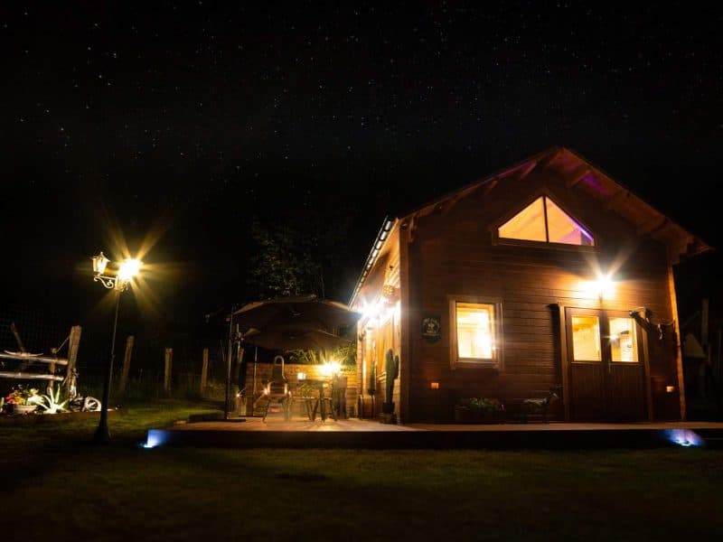 Chalet en bois illuminé sous un ciel étoilé, avec terrasse et mobilier extérieur.