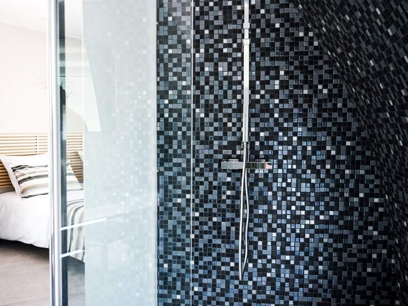 Chambre moderne avec douche à mosaïque noire et lit douillet en arrière-plan.