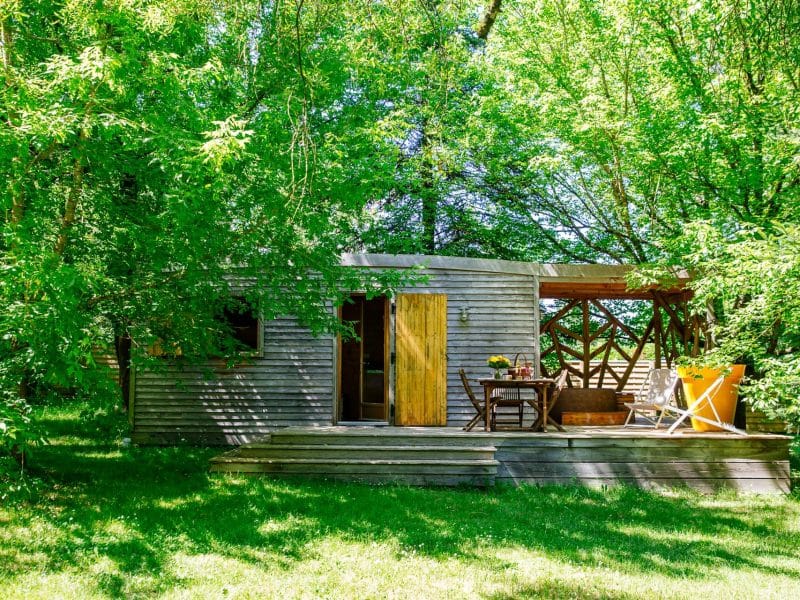 Cabane en bois nichée dans la verdure, avec terrasse ensoleillée et mobilier extérieur.