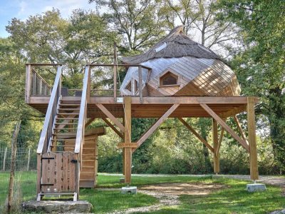 Cabane perchée en bois avec toit de chaume, entourée de verdure en Auvergne-Rhône-Alpes.