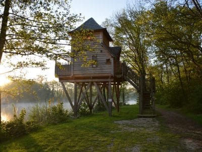Cabane perchée en bois sur pilotis, entourée de verdure au bord dun lac.