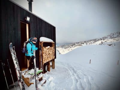 Cabane en bois dans la neige, avec skis à lentrée et vue sur les montagnes.