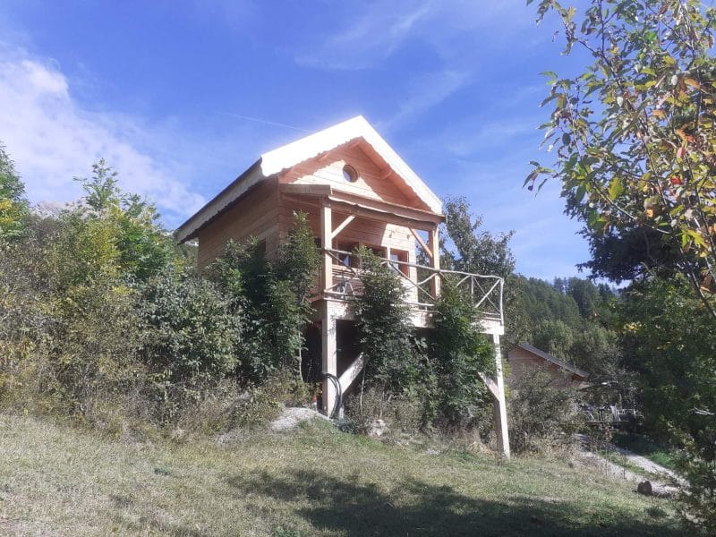 Chalet en bois avec balcon, entouré de verdure et sous un ciel bleu.