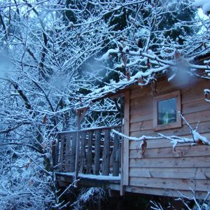 Cabane perchée en bois, entourée de neige et darbres enneigés. Tranquillité assurée.