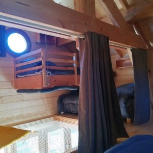 Cabane en bois avec un lit suspendu et une fenêtre ronde, ambiance chaleureuse.