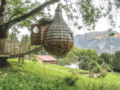 Cabane perchée en bois, suspendue à un arbre, avec vue sur les montagnes verdoyantes.