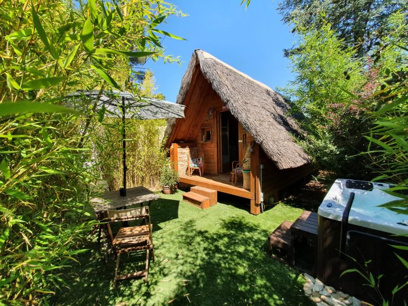 Chalet en bois avec toit de chaume, entouré de verdure et jacuzzi extérieur.
