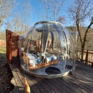 Hébergement insolite en bulle transparente, entouré darbres, avec terrasse en bois.
