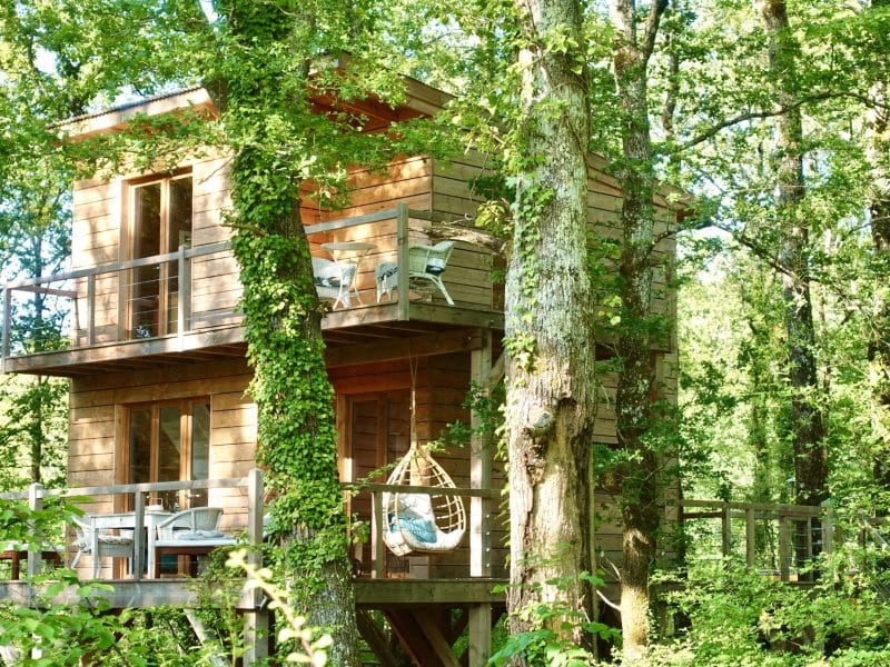 Cabane perchée en bois, entourée darbres, avec balcon et chaises confortables.