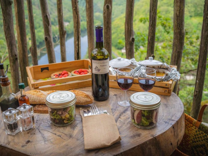 Table en bois avec un repas gourmet et du vin, vue sur la nature depuis un hébergement en hauteur.