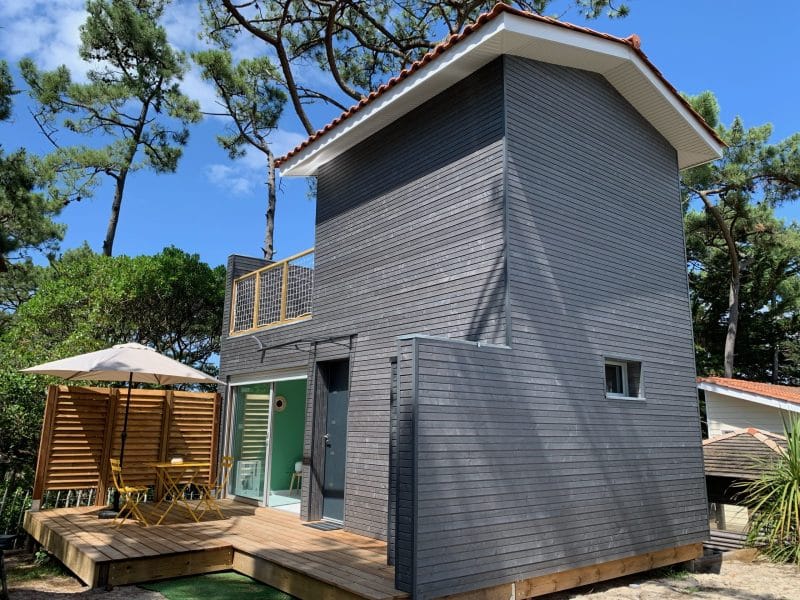 Maison moderne en bois avec terrasse, entourée de verdure et dun parasol.