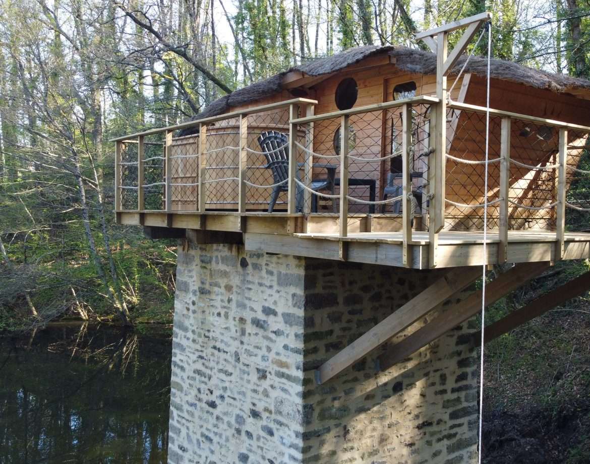 Cabane perchée sur pilotis, entourée de verdure, au bord dune rivière paisible.