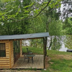 Cabane en bois au bord dun étang, entourée de verdure et dun coin salon extérieur.