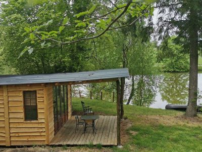 Cabane en bois au bord dun étang, entourée de verdure et dun coin salon extérieur.