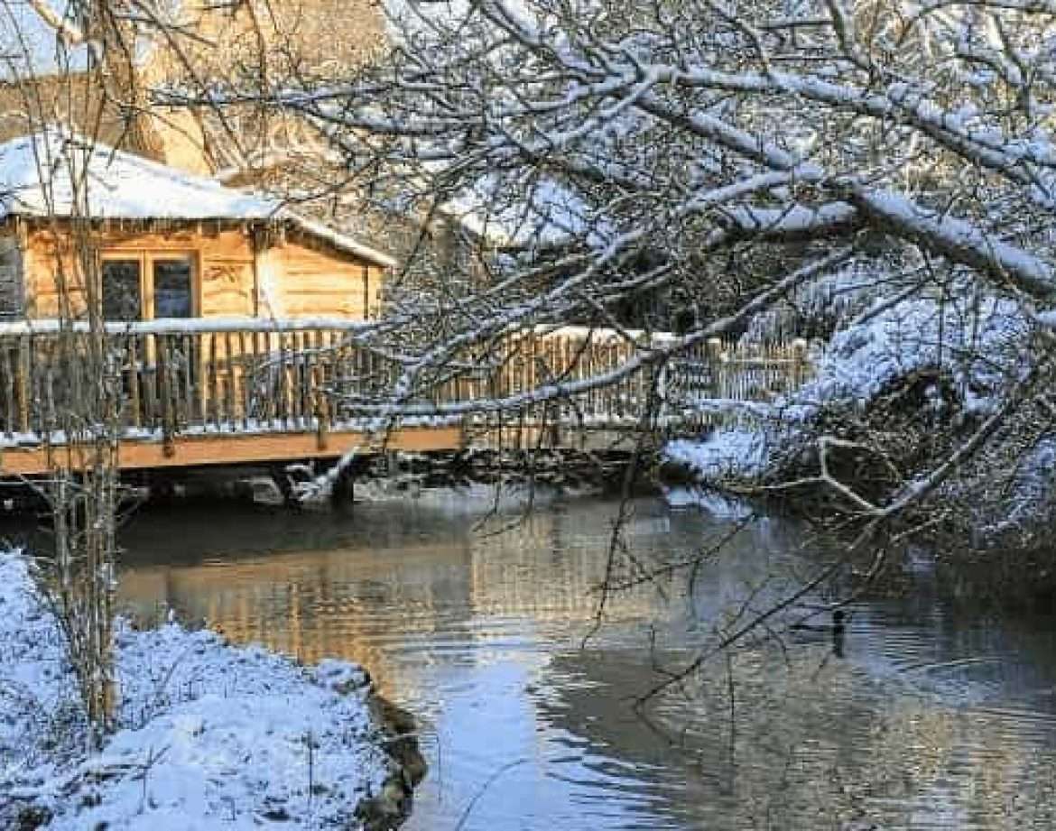 Cabane en bois au bord dun ruisseau, entourée de neige et de nature paisible.