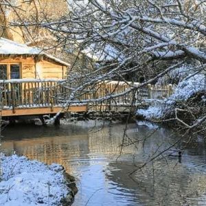 Cabane en bois au bord dun ruisseau, entourée de neige et de nature paisible.