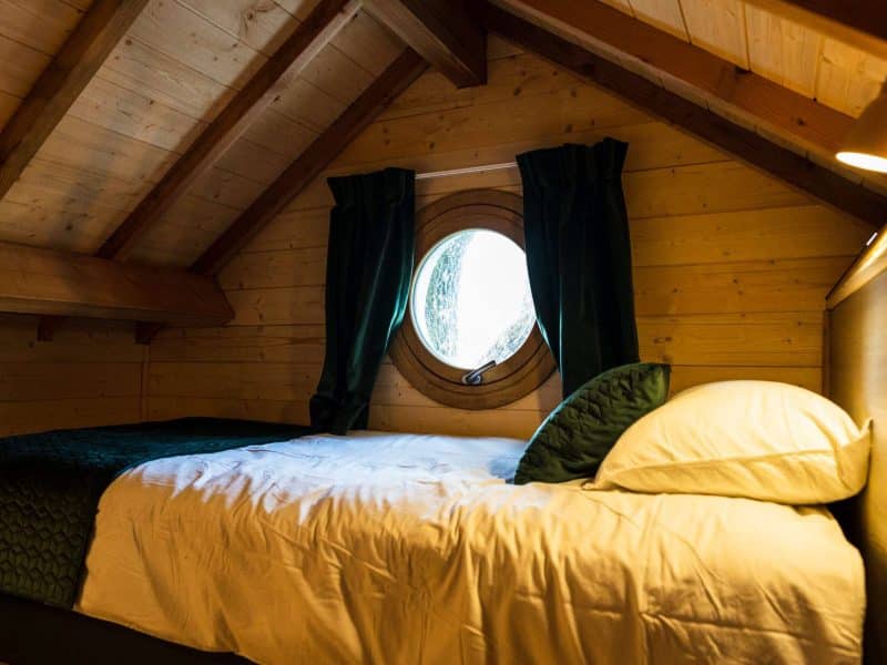 Chalet cosy avec lit confortable et fenêtre ronde, ambiance chaleureuse en bois.