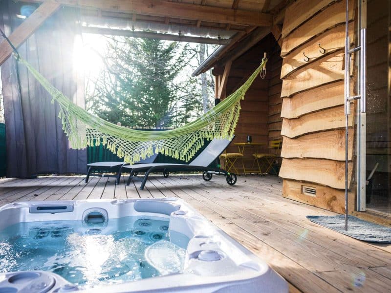 Cabane en bois avec jacuzzi, hamac suspendu et lumière naturelle apaisante.
