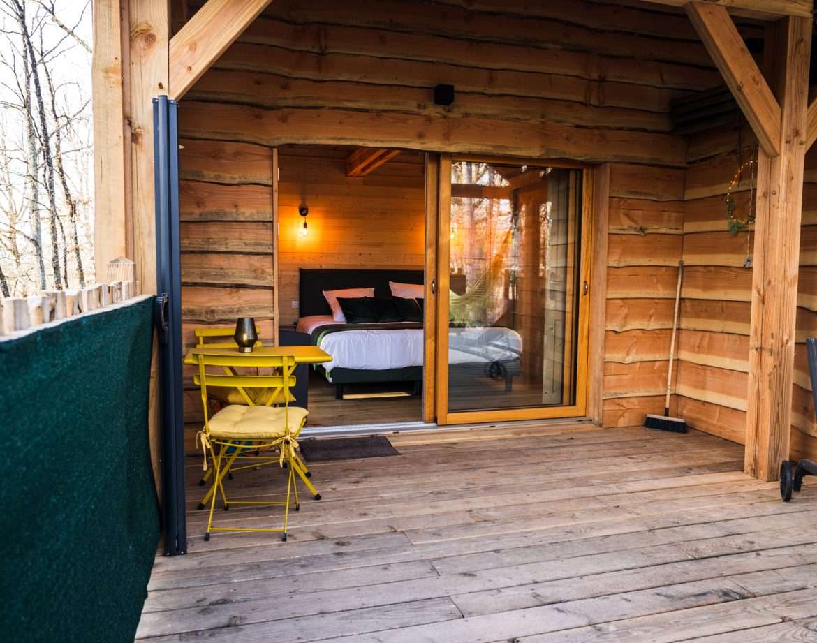 Cabane en bois avec terrasse, lit confortable et chaises jaunes accueillantes.