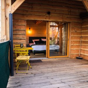 Cabane en bois avec terrasse, lit confortable et chaises jaunes accueillantes.