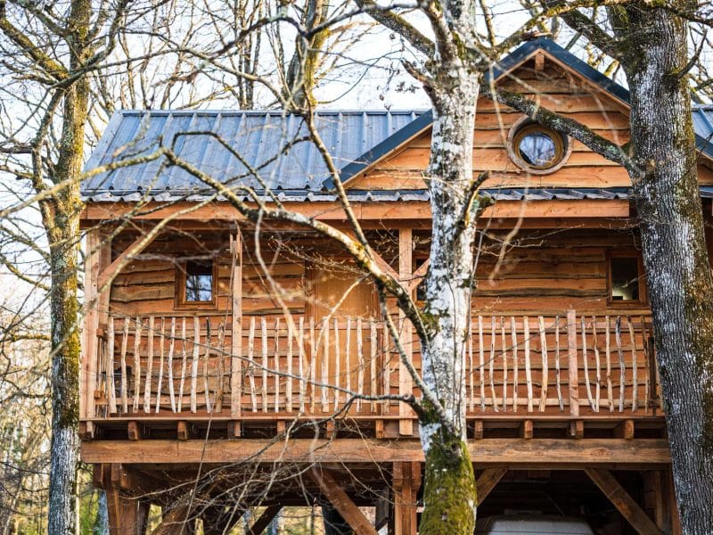 Cabane en bois perchée dans les arbres, avec un balcon en bois et un toit en métal.