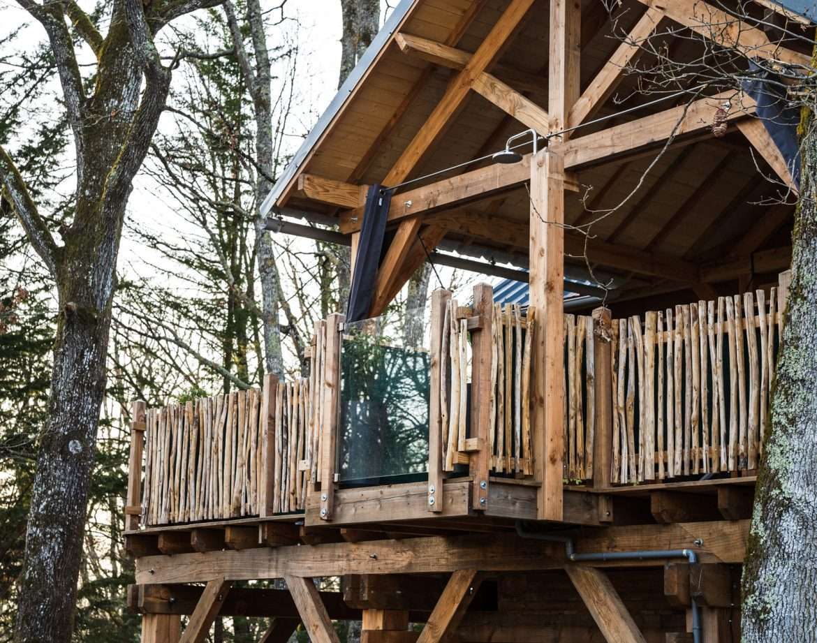 Cabane perchée en bois avec balcon, entourée darbres verdoyants.