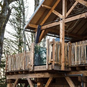 Cabane perchée en bois avec balcon, entourée darbres verdoyants.