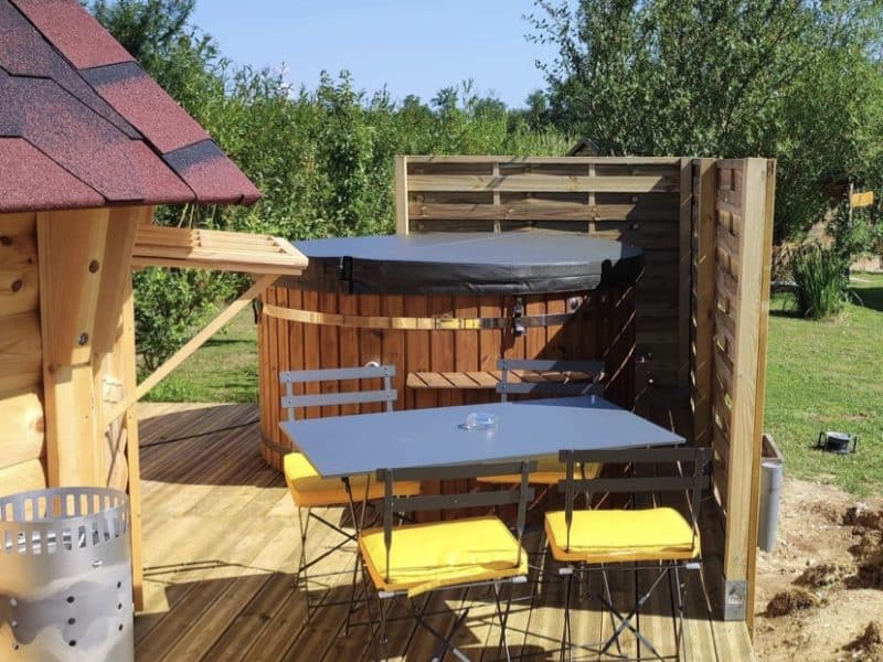 Chalet en bois avec terrasse, barbecue et chaises jaunes, entouré de verdure.