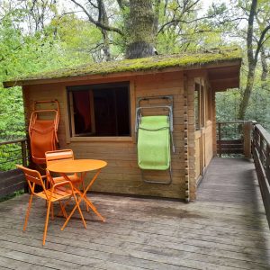 Cabane perchée en bois dans les arbres, avec terrasse et chaises colorées.