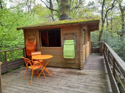 Cabane perchée en bois dans les arbres, avec terrasse et chaises colorées.