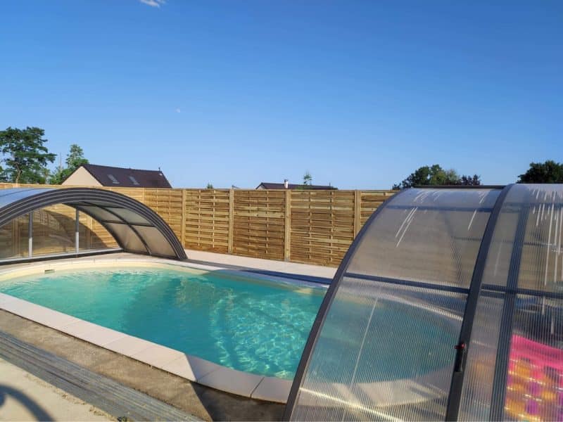 Villa avec piscine couverte, entourée dune clôture en bois et ciel dégagé.
