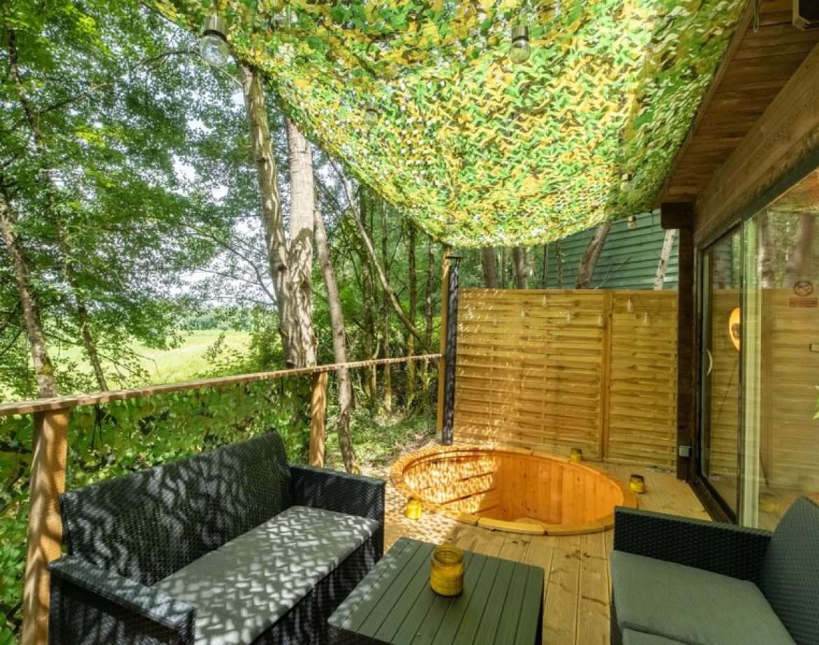 Cabane en bois avec terrasse ombragée et baignoire en bois, entourée de verdure.