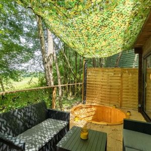 Cabane en bois avec terrasse ombragée et baignoire en bois, entourée de verdure.