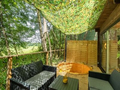 Cabane en bois avec terrasse ombragée et baignoire en bois, entourée de verdure.
