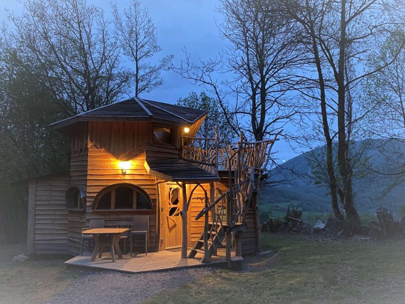 Cabane perchée en bois, illuminée au crépuscule, entourée darbres verdoyants.
