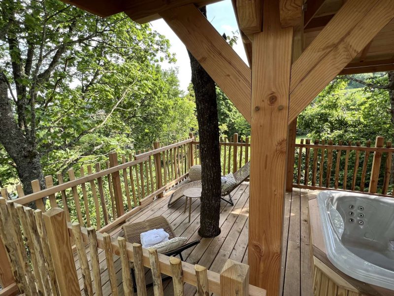 Cabane dans les arbres avec terrasse en bois et jacuzzi, entourée de verdure.