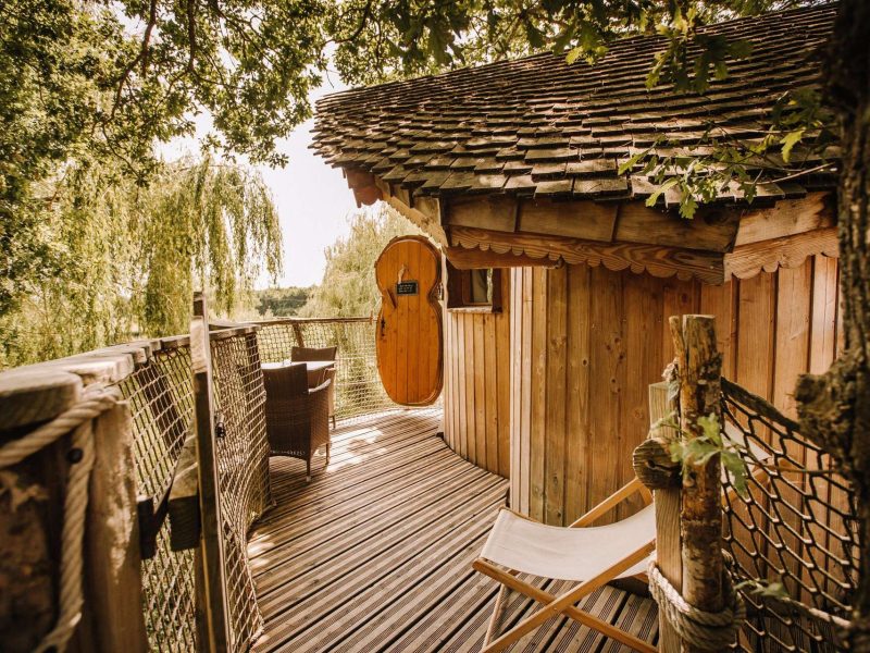 Cabane dans les arbres avec terrasse en bois et porte ronde, entourée de verdure.