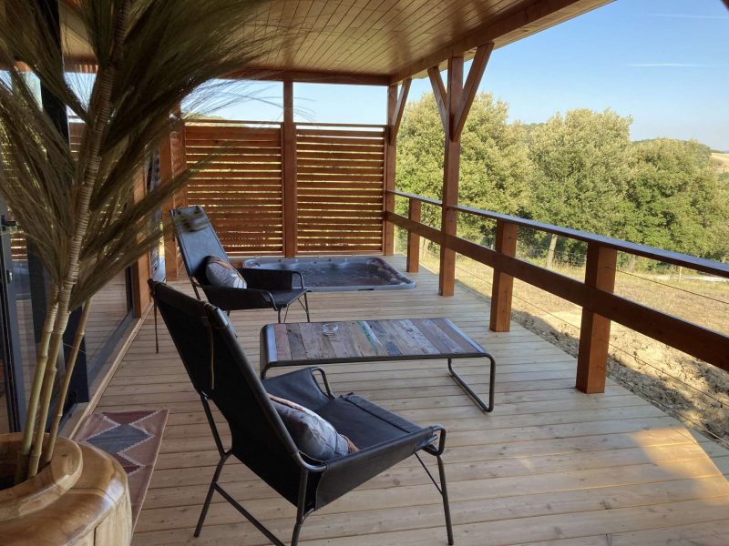 Chalet avec terrasse en bois, chaises confortables et vue sur la nature verdoyante.