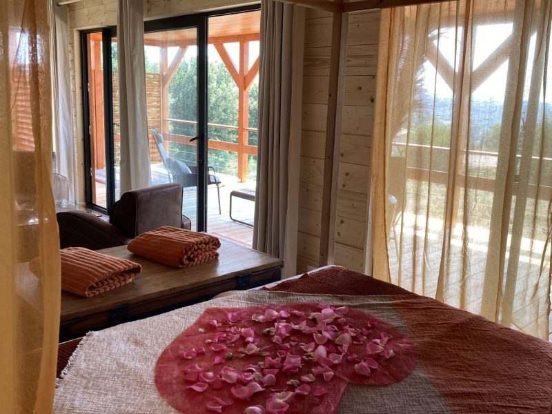 Chalet en bois avec vue, lit décoré de pétales de rose et balcon ensoleillé.