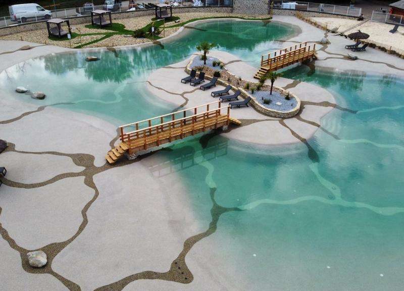 Hébergement de luxe avec piscine naturelle et pont en bois, entouré de chaises longues.