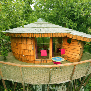 Cabane en bois perchée, entourée darbres, avec terrasse et mobilier coloré.