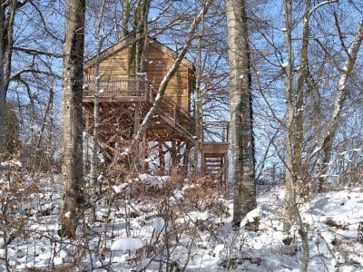 Cabane perchée en bois dans les arbres, entourée de neige et de nature.