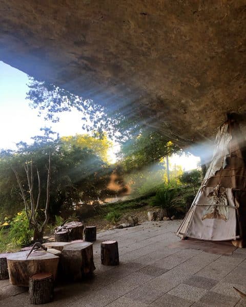 Tente de style tipie sous un rocher, entourée de nature verdoyante et de troncs en bois.