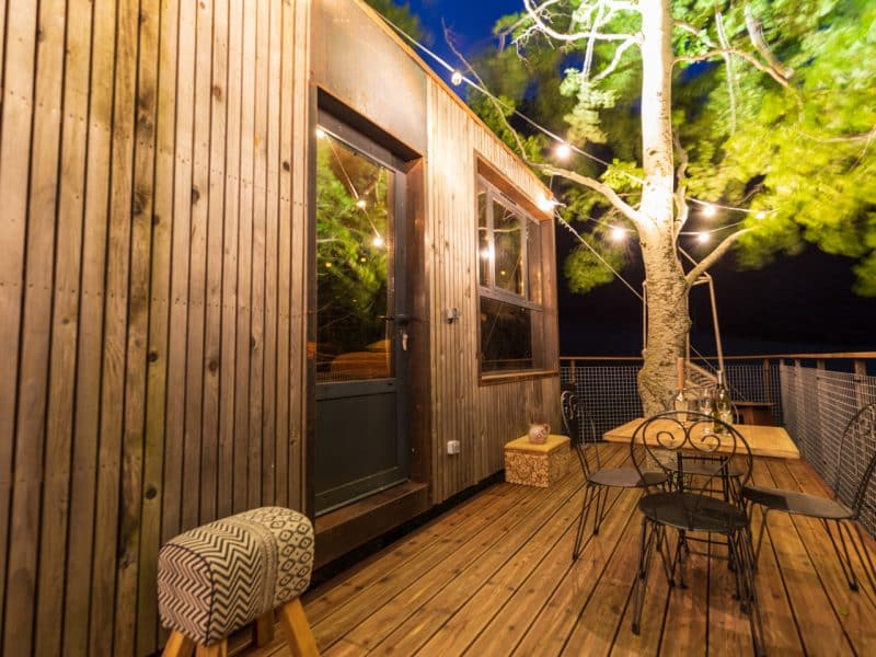 Cabane en bois avec terrasse illuminée, entourée darbres. Ambiance chaleureuse.