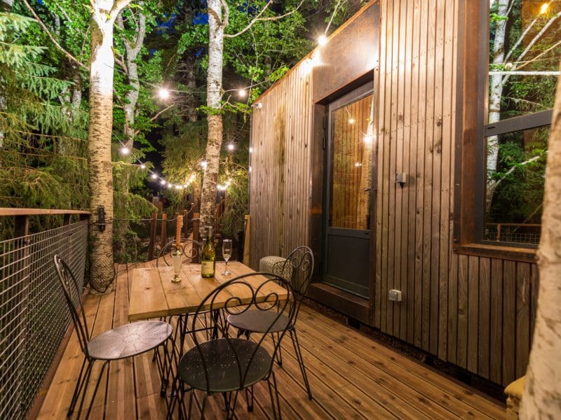 Cabane dans les arbres avec terrasse en bois, éclairage féérique et vue sur la forêt.