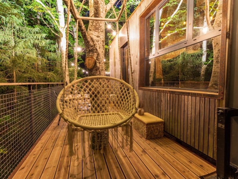 Cabane perchée dans les arbres avec un fauteuil suspendu et des lumières douces.