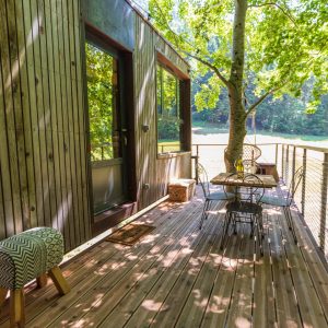 Cabane perchée en bois avec terrasse ombragée par un arbre, vue sur la nature.