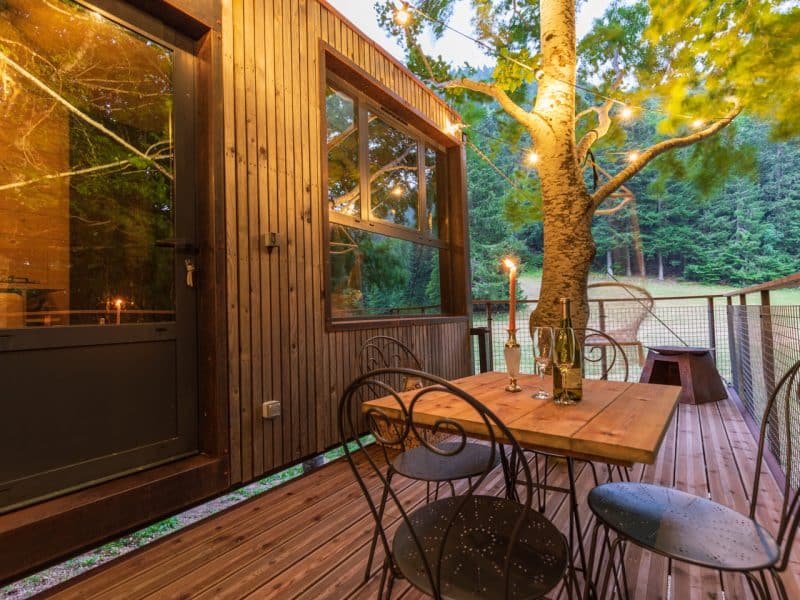 Cabane en bois avec terrasse, éclairée par des guirlandes, entourée darbres.