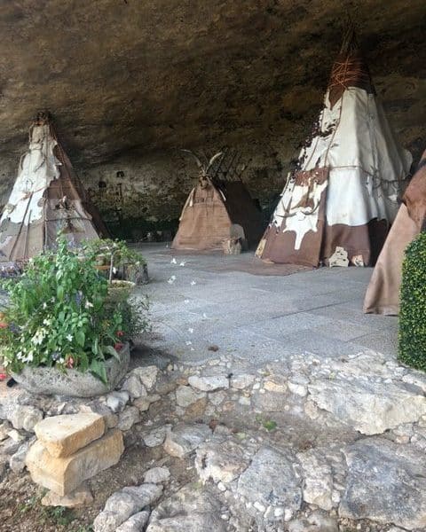 Tipis traditionnels sous une roche, entourés de verdure et de pierres.